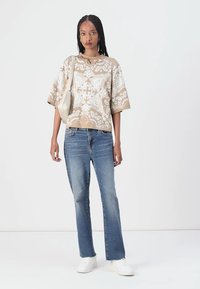 Beige mønstrete T-skjorte med korte ermer kombinert med lyseblå straight-leg jeans og hvite sneakers. Tilbehøret inkluderer en lys veske.
