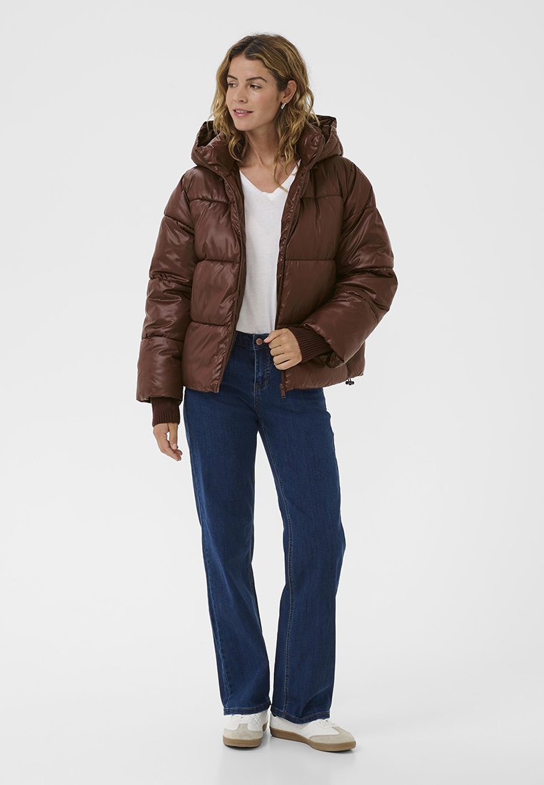 Braune Pufferjacke mit Kapuze, glänzender Oberfläche, gestepptem Design und gerippten Bündchen, kombiniert mit blauen Jeans und weißen Schuhen.