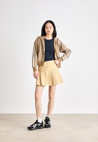 Bruine geblokte zipjack, marineblauw shirt, gele plooienrok en zwarte sneakers met witte accenten en dikke zolen. Casual outfit setting.