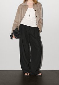 Beige uldjakke med sorte knapper, hvid tanktop, sorte brede bukser og sorte flip-flops; holder en vævet sort håndtaske.