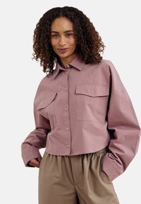 Grezni, gaiši mauve krāsas kokvilnas krekliņš ar lielām krūšu kabatām, brīvs piegriezums un garas piedurknes ar platiem aprocēm. Apvienots ar brīviem bēšiem biksēm.