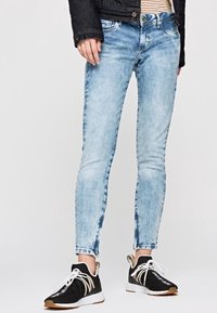 Personne portant un jean skinny bleu clair, des baskets noires et blanches, une chemise rayée et une veste sombre, se tenant contre un fond blanc.