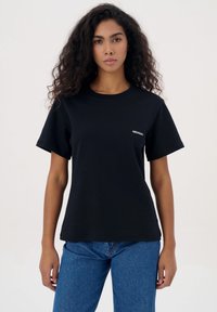 Zwarte katoenen t-shirt met korte mouwen, ronde hals en klein wit "MISAGA" logo op de linkerkant van de borst. Gekocht met blauwe jeans.