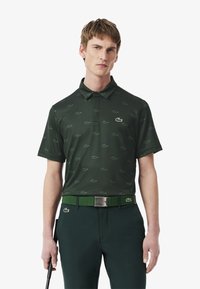 Mörkgrön polo med ett repetitivt mönster av ljusgröna krokodiler. Har en krage och korta ärmar. Matchas med ett grönt bälte.