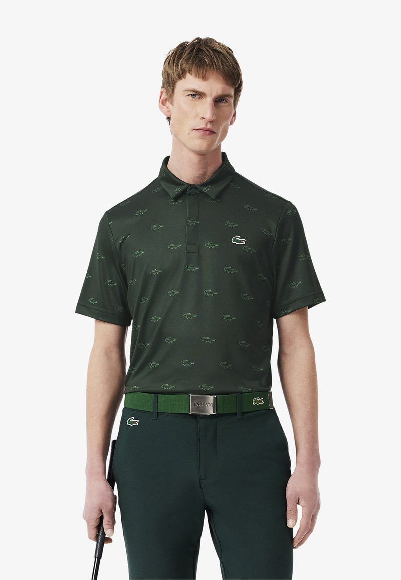 Mörkgrön polo med ett repetitivt mönster av ljusgröna krokodiler. Har en krage och korta ärmar. Matchas med ett grönt bälte.