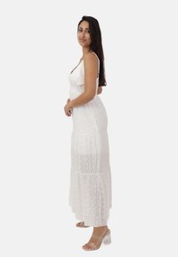 Witte, mouwloze maxi jurk met perforaties, een gelaagde rok en een aansluitend lijfje. Gecombineerd met doorzichtige sandalen met hak.