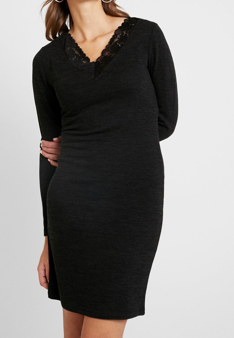 Robe noire à manches longues avec un décolleté en V orné de dentelle. Le tissu semble doux et extensible, épousant étroitement le corps.