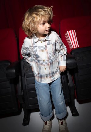 Bambino in una camicia a quadri bianca con bottoni dorati, jeans blu con risvolto, e sneakers bianche, in piedi tra i posti del cinema.