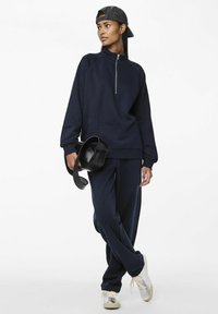 Felpa navy con zip e maniche raglan, abbinata a pantaloni della tuta tapered coordinati. Una borsa a tracolla nera e sneakers metalliche completano il look.