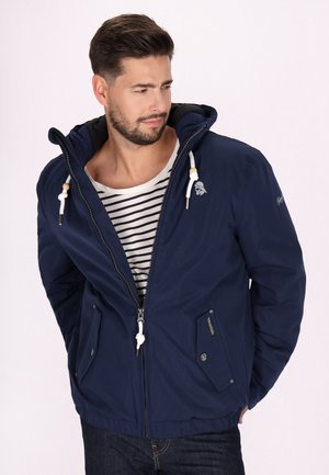 Marineblaue Kapuzenjacke mit Frontreißverschluss, mit Zugbandakzenten und zwei Seitentaschen. Über einem schwarz-weißen gestreiften Shirt getragen.