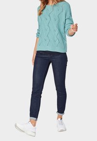 Pull tricoté bleu clair avec un motif en forme de vague, associé à un jean foncé et des chaussures de sport blanches. Le pull est ample avec des manches retroussées.
