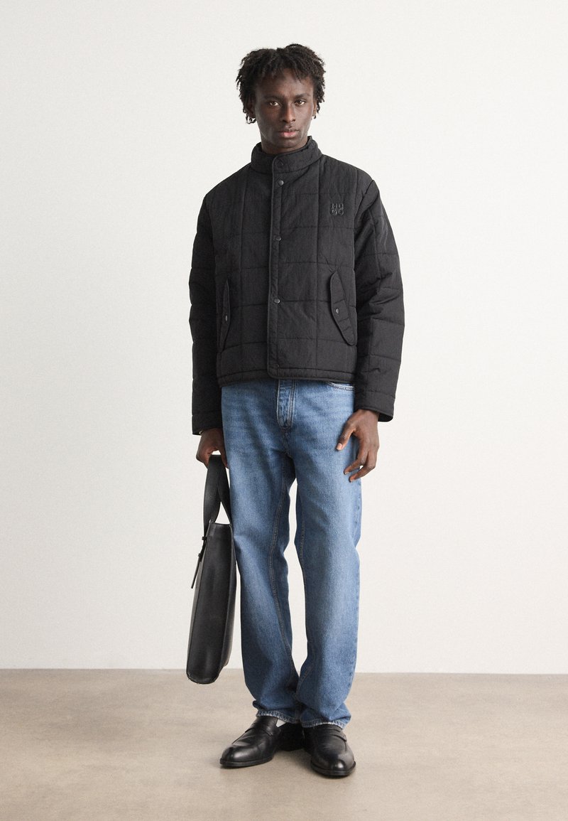 Veste noire matelassée avec col haut, dotée de boutons-pression et de poches latérales. Associée à un jean bleu clair et des chaussures noires, portant un sac noir.