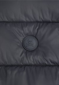 La section de la veste matelassée noire présente une finition mate, avec un patch en caoutchouc noir rond arborant un emblème de crâne et os croisés.