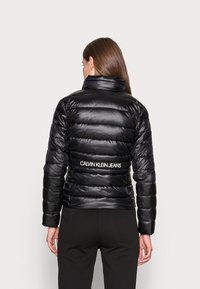 Kobieta o długich brązowych włosach, ubrana w czarną pikowaną kurtkę z napisem "Calvin Klein Jeans" na plecach, w połączeniu z czarnymi spodniami.