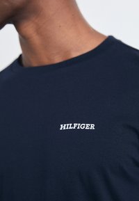 Marineblå bomulds T-shirt med et hvidt "HILFIGER" logo på brystet, rund hals og glat tekstur. Korte ærmer og afslappet pasform.