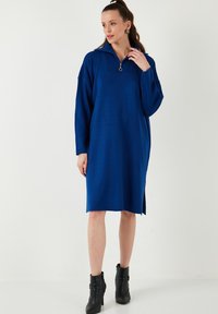 Rochie albastră cu mânecă lungă, cu guler cu fermoar, croi lejer, până la genunchi și slituri laterale, fabricată dintr-un material moale; asortată cu cizme negre până la glezna.