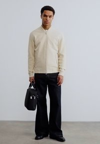 Νέος άνδρας με μπεζ τζάκετ με φερμουάρ και μαύρο παντελόνι φαρδύ, κρατά μια μαύρη τσάντα εργασίας, στέκεται μπροστά σε έναν απλό λευκό τοίχο.