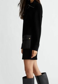 Schwarzes geripptes Strickkleid mit hohem Kragen, ausgestattet mit einer großen schwarzen Tasche mit Druckknöpfen. Kombiniert mit strukturierten schwarzen kniehohen Stiefeln.