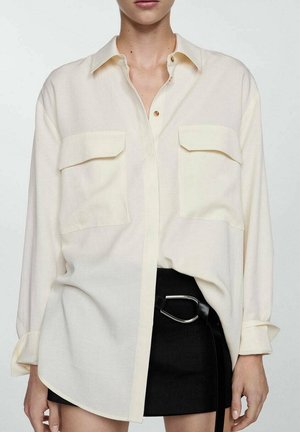 Femme portant une chemise crème oversize boutonnée avec deux poches avant et une mini-jupe noire avec une ceinture à boucle argentée.