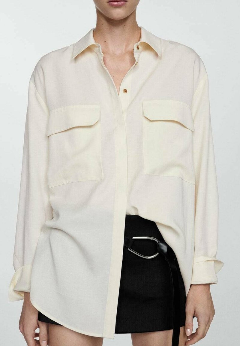 Femme portant une chemise crème oversize boutonnée avec deux poches avant et une mini-jupe noire avec une ceinture à boucle argentée.