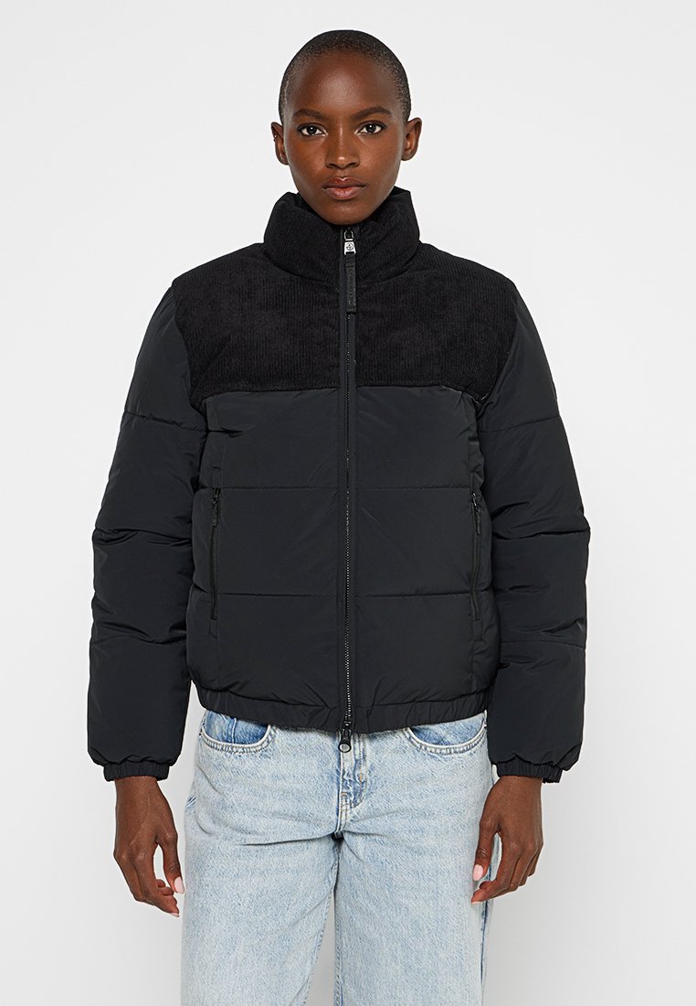 Person, der eine schwarze Steppjacke mit strukturiertem Oberteil und hellblauen Jeans trägt, steht mit entspannten Armen an den Seiten.