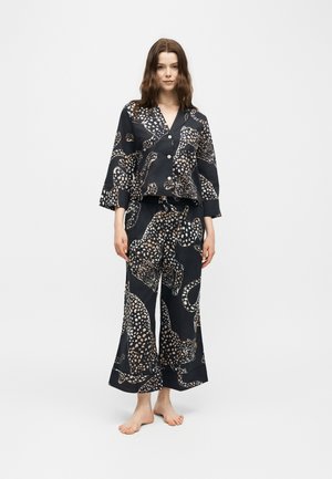 Jonge vrouw staat blootsvoets en draagt een zwart bijpassend pyjamaset met een beige en witte luipaardprint op een effen achtergrond.