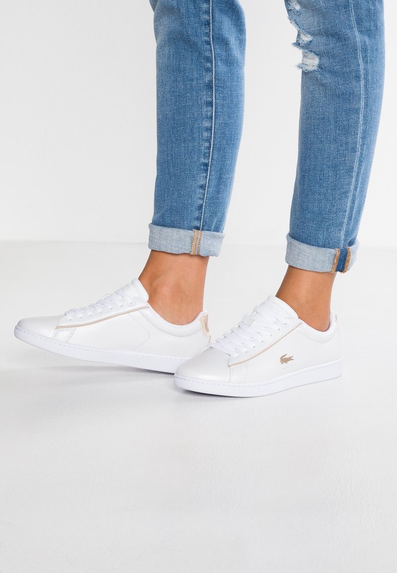 Personne portant un jean bleu retroussé et des baskets basses blanches avec des accents beiges, debout sur une surface blanche.