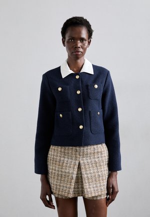 Navy textured jacket with gold buttons and four front pockets, paired with a plaid tweed skirt. White shirt collar adds contrast.