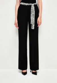 Zwarte wijde broek met een zachte textuur, voorzien van een contrasterende panterprint sjerp die om de taille is geknoopt, aangevuld met puntige zwarte hakken.