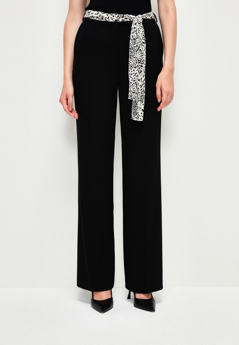 Zwarte wijde broek met een zachte textuur, voorzien van een contrasterende panterprint sjerp die om de taille is geknoopt, aangevuld met puntige zwarte hakken.