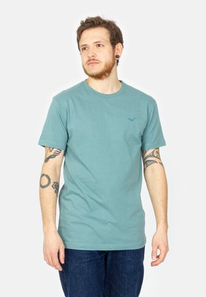 Türkise Baumwoll-T-Shirt mit kurzen Ärmeln und Rundhalsausschnitt. Auf der linken Brust befindet sich ein kleines blaues Logo, und es wird mit dunklen Jeans kombiniert.