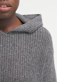 Grijze, gestructureerde gebreide hoodie met een opvallende capuchon. Het ontwerp heeft een losse pasvorm en zichtbare stiksels, wat een gezellige uitstraling creëert.