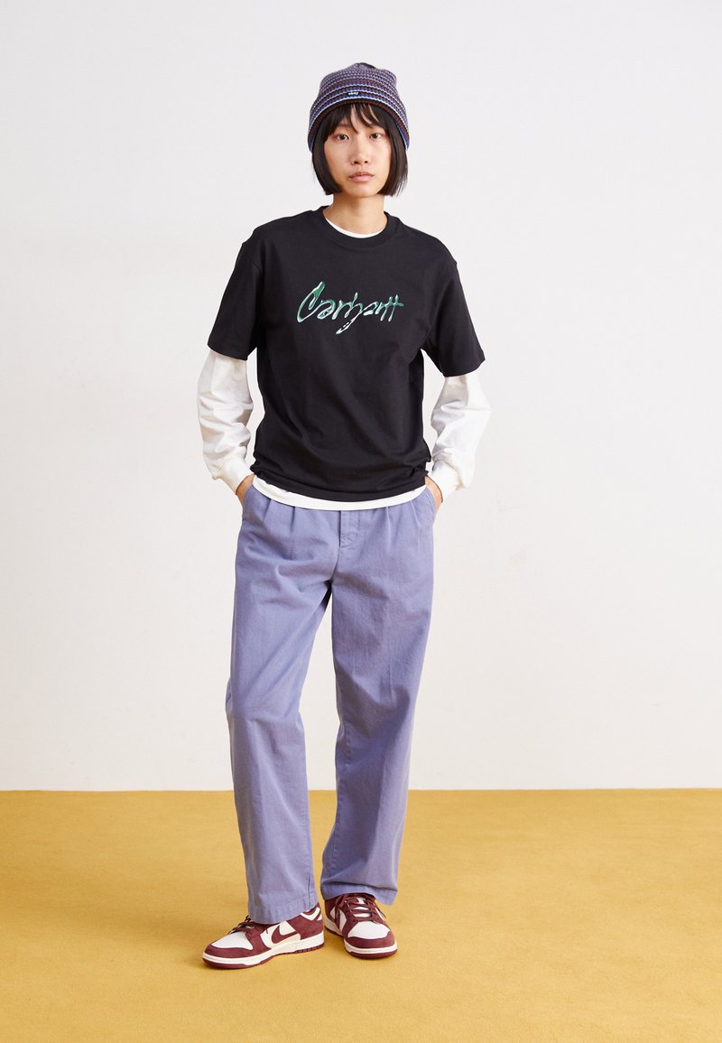 Camiseta de algodón negra con logo "Carhartt" en turquesa, superpuesta a una camisa de manga larga blanca. Pantalones de pierna ancha azul claro, zapatillas rojas y blancas.