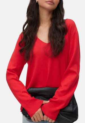 Donna che indossa un maglione a maniche lunghe a V di colore rosso acceso, con una borsa clutch nera davanti a uno sfondo chiaro.