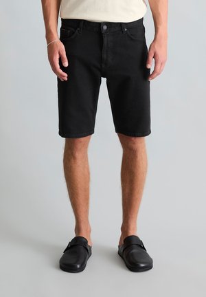 Man die zwarte knielange shorts draagt, beige overhemd, zwarte instapschoenen en een zilveren armband, staand op een grijze achtergrond.