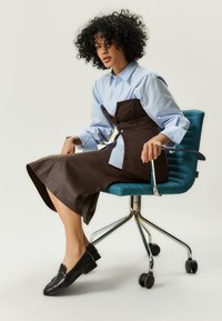 Abito corsetto marrone sovrapposto a una camicia azzurro chiaro, abbinato a scarpe slip-on nere. Seduto su una sedia da ufficio teal con base metallica.