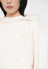 Detajlni pogled na osebo, oblečeno v svetlo roza kapuco z teksturiranim dizajnom mašne blizu levega rame in vidnim sprednjim žepom.