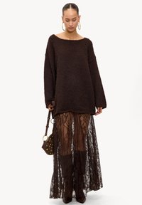 Vrouw in een oversized fuzzy bruin trui, doorzichtige zwarte kanten maxi rok, bruine laarzen, grote hoepel oorbellen en met een bruin gestikt handtas in de hand.