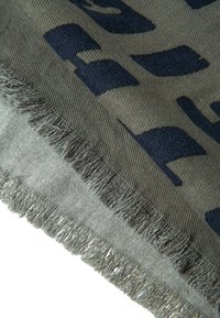 Knit scarf in olive green with navy abstract pattern. Features a fringed edge and varied textures, highlighting contrasting color layers.