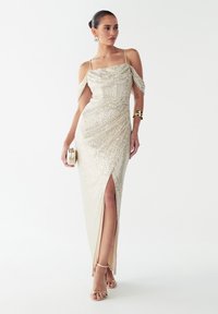 Večerna obleka v kovinsko zlati barvi z biseri, s prilagojeno silhueto, raglanskimi rokavi in visokim razporkom do stegna. Doplačana z zlatimi dodatki.