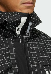 Musta takki, jossa on ruutukuosi, korkea kaulus ja vetoketjusuljenta. Materiaali näyttää sileältä ja siinä on hienovarainen kiilto, ja se esittelee brändättyjä yksityiskohtia.