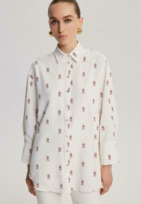 Donna che indossa una camicia bianca con bottoni decorata con un piccolo motivo floreale rosso e grandi orecchini a cerchio dorati, su uno sfondo neutro.