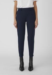 Pantalons ajustés navy sur mesure, en tissu lisse ; dotés de poches avant et d'une ceinture plate. Associés à des bottines noires.