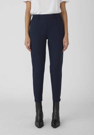 Pantalons ajustés navy sur mesure, en tissu lisse ; dotés de poches avant et d'une ceinture plate. Associés à des bottines noires.