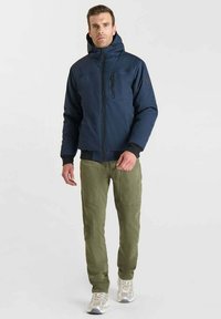 Giacca isolante blu navy con cappuccio, tasca sul petto con zip e polsini a coste neri, abbinata a pantaloni verde oliva e sneakers chiare.