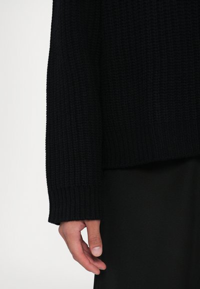 Primo piano di una mano che pende rilassata accanto a un maglione nero a maniche lunghe in maglia a coste e pantaloni neri su uno sfondo semplice.