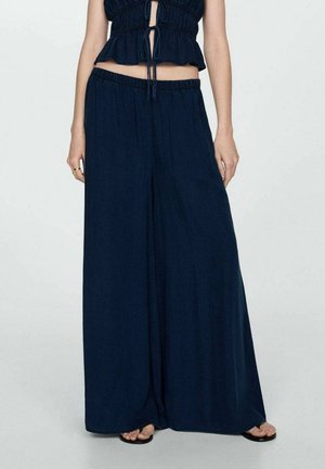 Femme portant un pantalon à jambes larges bleu foncé avec une taille élastique et des sandales noires, associé à un haut sans manches bleu foncé avec des liens.