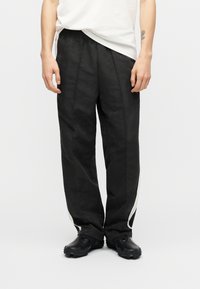 Άτομο που φοράει φαρδιά μαύρη παντελόνια με λευκές ρίγες στο πλάι και μαύρα παπούτσια με υφασμένο πάτο, στέκεται σε απλό φόντο.