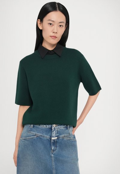 Īsroku krekls tumši zaļā krāsā ar gludu tekstūru un apaļu kakla izgriezumu, uzvilkts virs melnas apkaklītes krekla, kopā ar džinsa svārkiem.