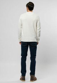 Witte blouse met lange mouwen en ribbelstructuur, gecombineerd met donkerblauwe jeans. Bruine laarzen met zichtbare veters en robuuste zolen. Verstelbare mouwen.